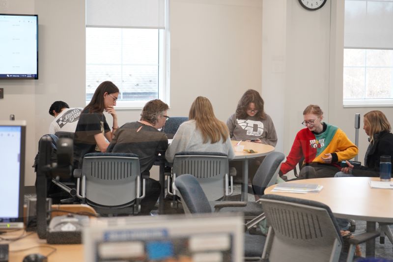 students, a librarian and their professor sit at a round table in the library classroom using their laptops for research