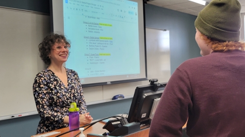 Image of smiling Program Director, Dr. Jamie Small on the left side on the screen behind a classroom podium. On the right side of the screen is a student speaking to Dr. Small with their back to the camera.