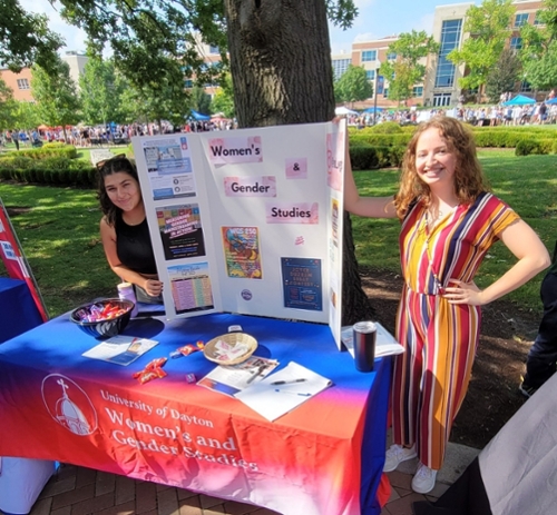 Image of two smiling women standing on either side of a table holding a Poster Board. The poster board says Women's and Gender Studies and includes several flyers. The table is covered by a blue and red tablecloth, the front reads University of Dayton, Women's and Gender Studies. There is a bowl of candy on the table. The table is under a tree, behind is a field with a crowd of students. The photograph was taken at the event "Up the Orgs". 