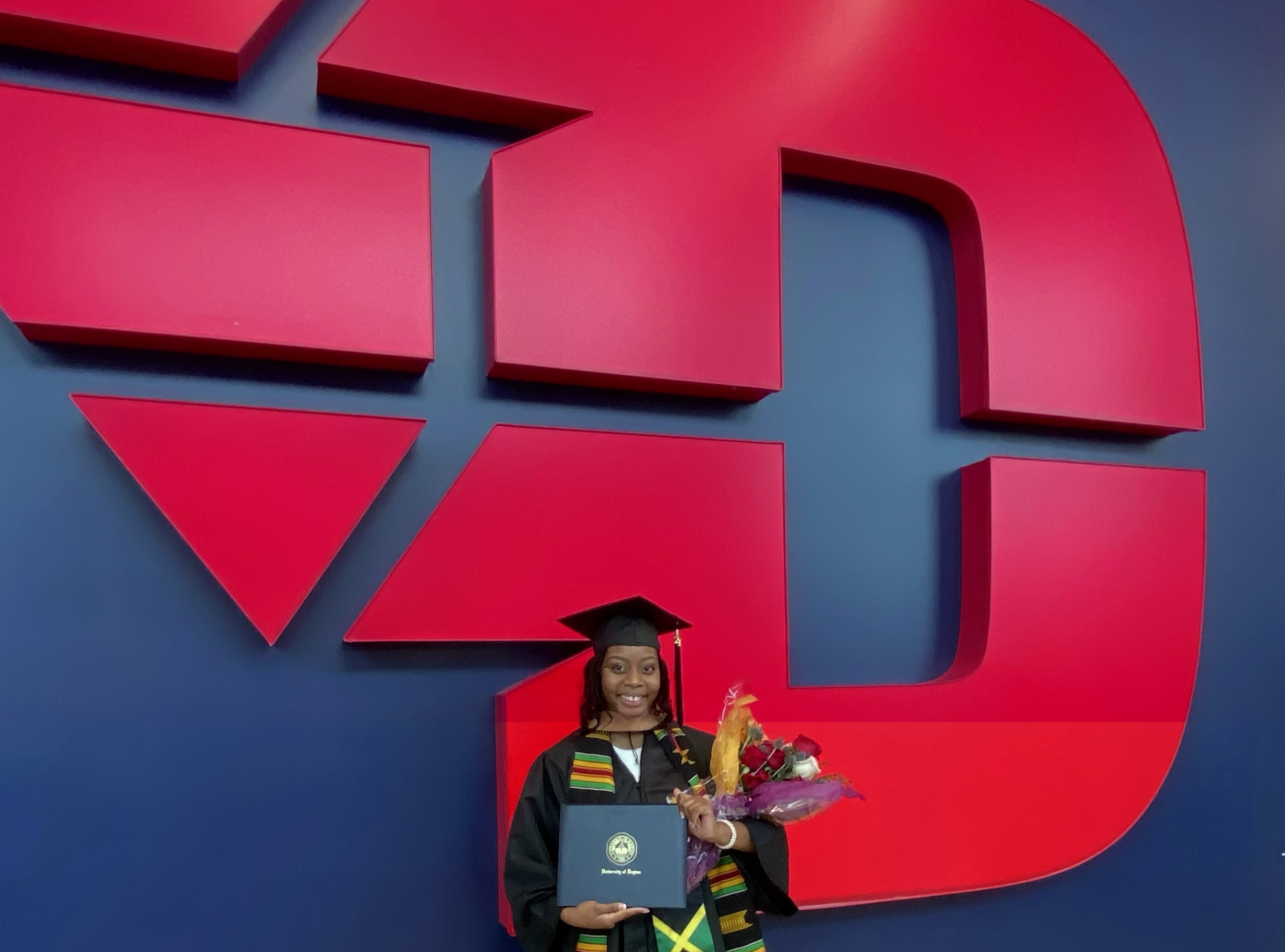 Danielle posing in front of the UD logo at UD arena in her cap and gown after graduating