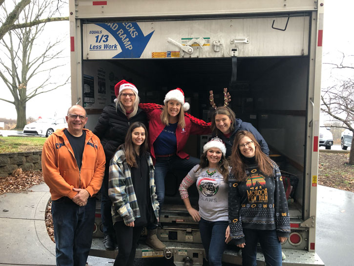 Seven adults pose in front of the back of a moving truck. They are decked out in holiday sweaters and festive hats.