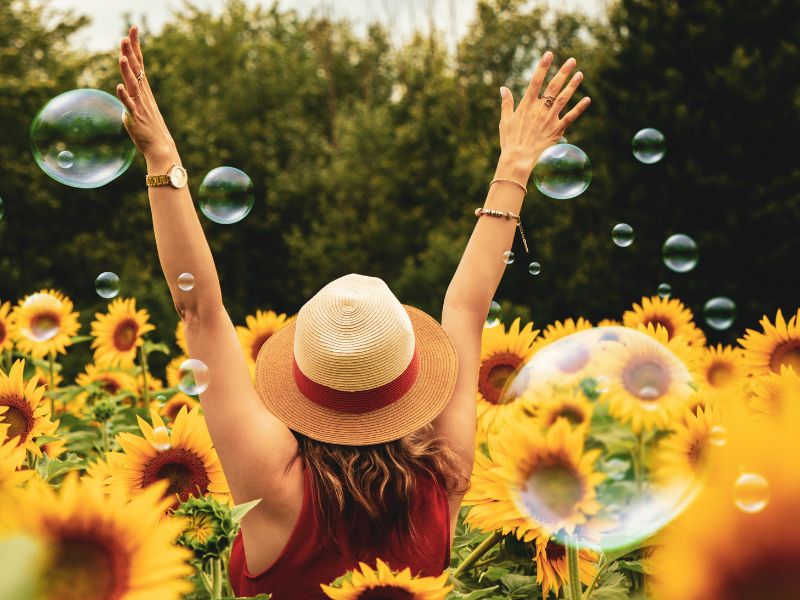 Photo of the back of a woman standing in a field of sunflowers with her arms outstretched. There are bubbles in the air.