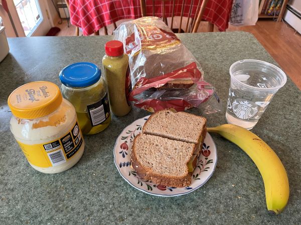 A sandwich on a ceramic plate, surrounded by a banana, a plastic Starbucks cup of water, a bag of bread, a mustard bottle, a jar of pickles, and a container of mayonnaise.