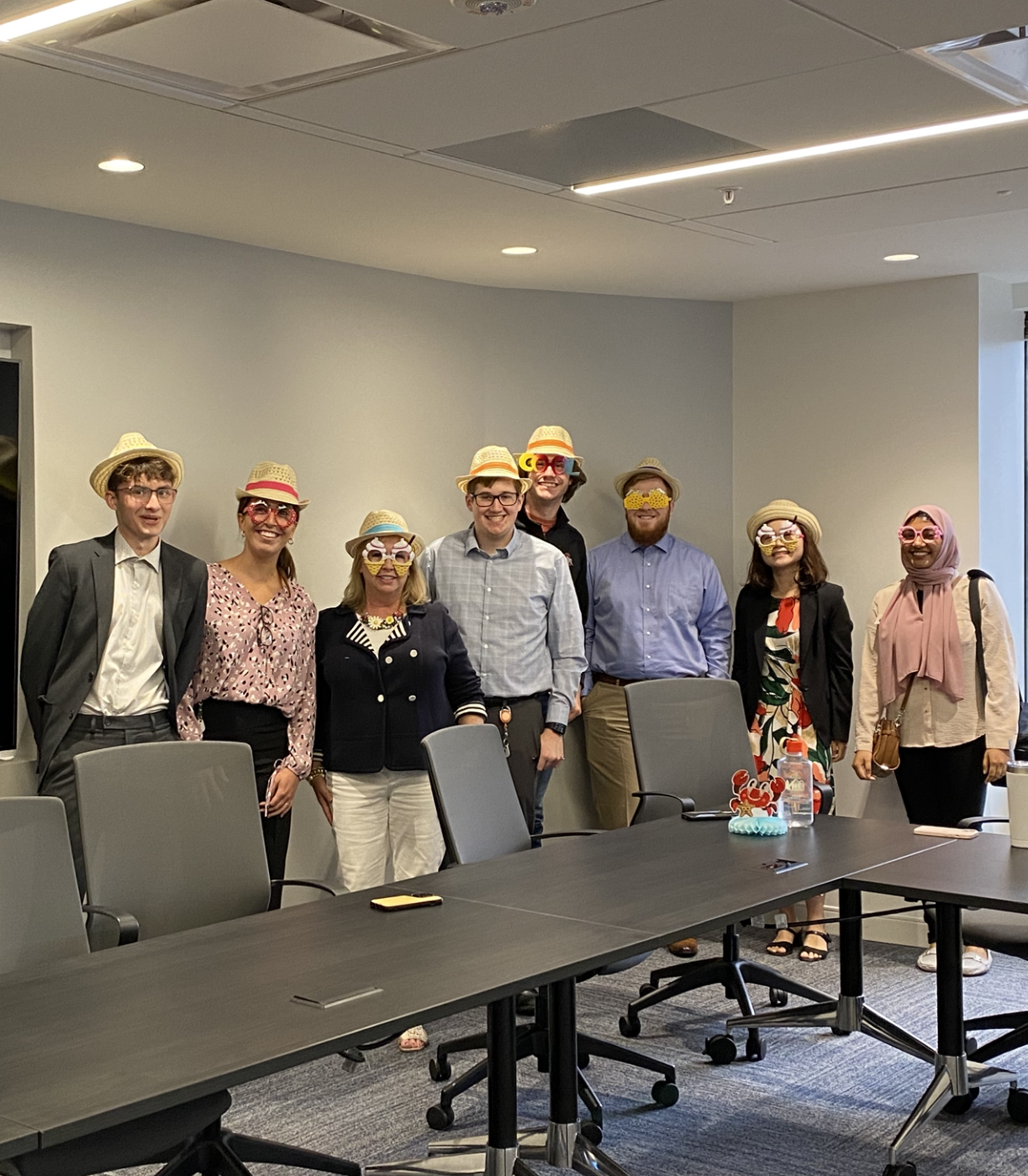 Eight people wearing fedoras and funny glasses during a work event. 
