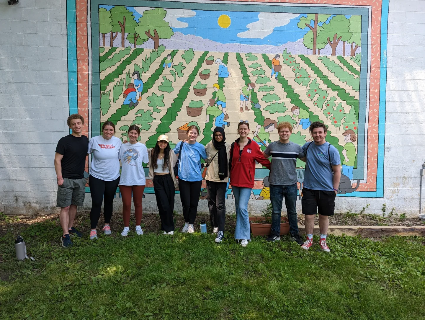 Nine students standing in front of a mural of farm while volunteering at a community farm. 