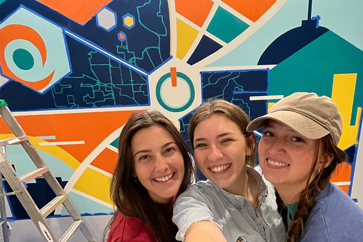 Gracie, Claire and Mira standing in front of their work in progress, the entryway mural of The Hub Powered by PNC Bank. To the left, there is a ladder; the background is the mural mostly finished painting, but still covered in painter's tape; and the three alumni are in the bottom right corner, smiling.