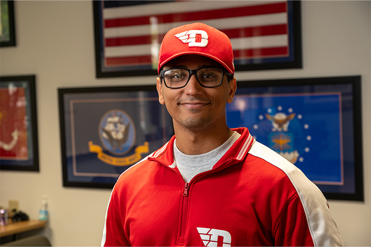 Shelby Dennhardt, wearing a red Dayton Flyers shirt and hat, standing in the Military and Veteran Programs and Services Center.