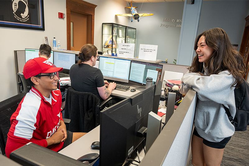 Shelby Dennhardt seated behind a counter with two other student workers in the Military and Veteran Programs and Services Center, speaking with another student to the right of him over the counter.