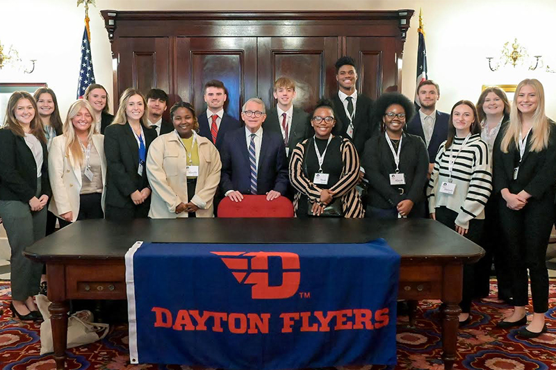 2023 Dayton2Columbus group with Ohio Gov. Mike DeWine in his office, holding a UD flag.