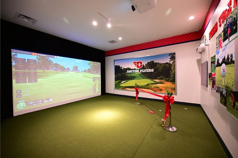 Inside the NCR Player Development Center featuring a large screen on the far wall and putting green. There is a red ribbon in celebration of the facility opening.