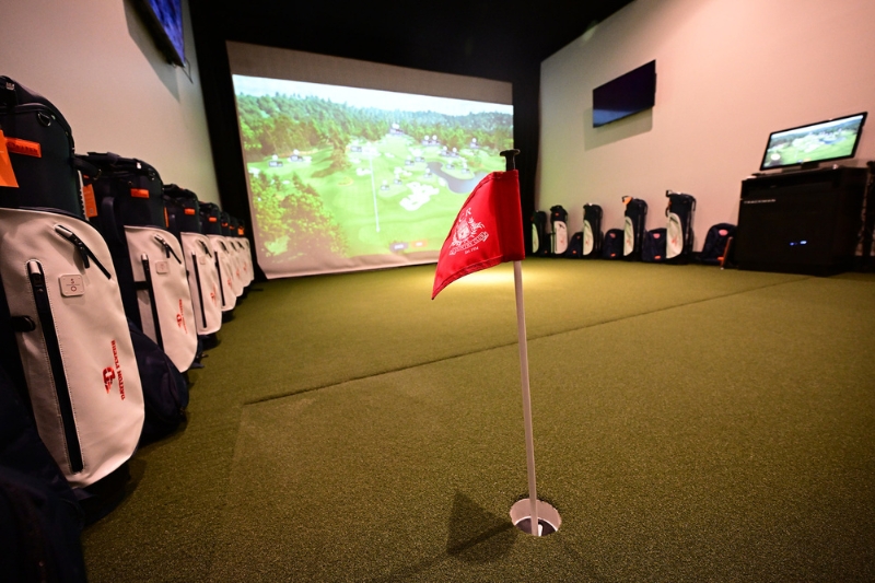 The NCR Player Development Center with a large monitor and putting green. There are golf bags lined up along the left-side wall.
