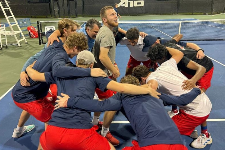 Men's tennis team in a circle with their arms around each other, and slightly crouched. One player is standing in the center.