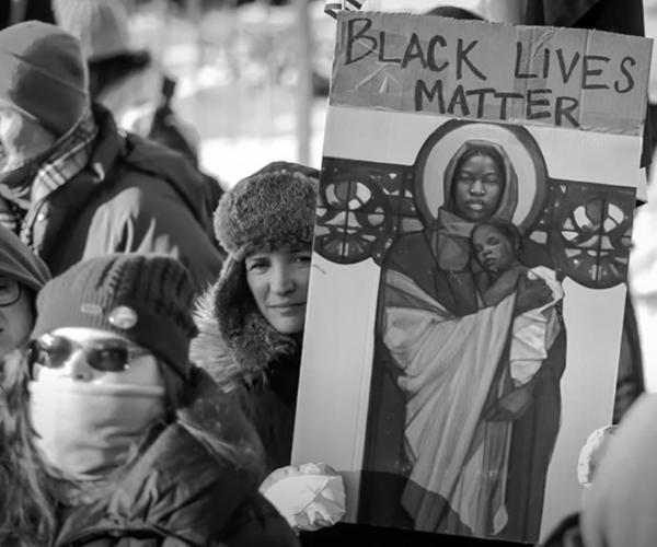 This image depicts a demonstrator holding a reproduction of McKenzie’s painting “Mary and Jesus with the Papel Picado” with the Black Lives Matter slogan