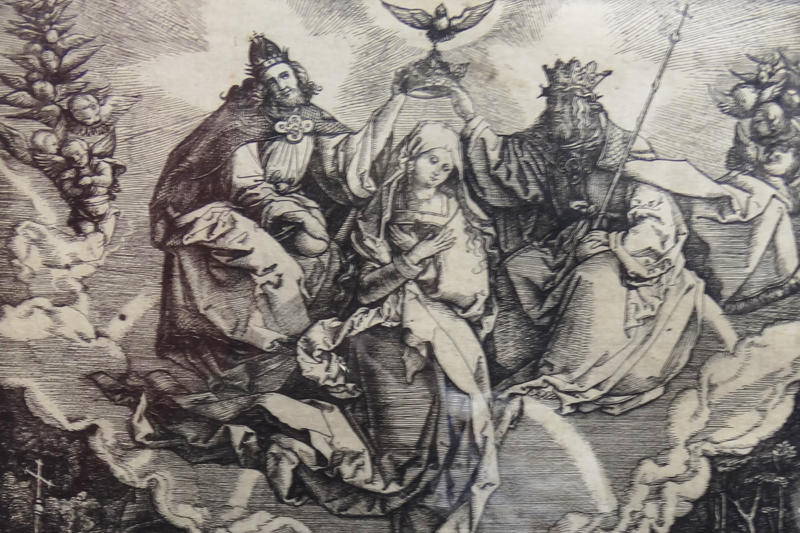 Black and white print. The Blessed Virgin Mary, sitting, is centered on the top half of the image as if rising. Her head is bowed, and her arms are crossed at her chest. A dove representing the Holy Spirit is above her head with a depiction of God the Father on the right and Jesus on the left, each holding the crown above Mary’s head. A grouping of angels is on each side.