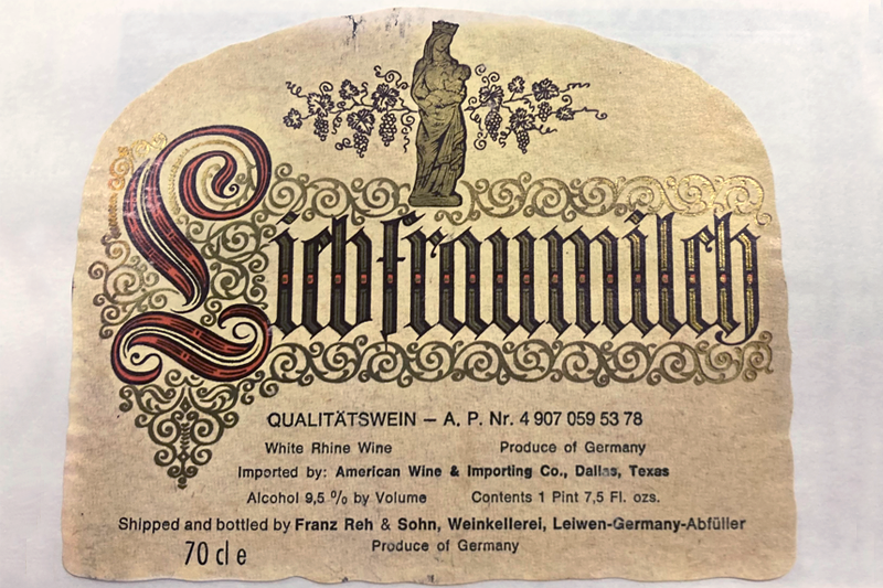 Label removed from a bottle of “Liebfraumilch white Rhine wine produced in Germany. Shipped and bottled by Franz Reh &amp; Sohn, Weinkellerei, Leiwen-Germany-Abfüller.” The label is a light golden paper, rounded at the top. A gold, outlined in black, standing Madonna nursing the Infant Jesus in the middle of illustrated grapevines is at the top above “Liebfraumilch” which is set in an old style script. The “L” is red and the rest of the letters are gold. “Liebraumilch” is surrounded by gold scrolls.