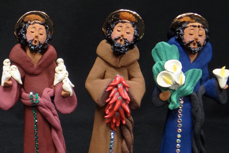 Three clay figures of men in robes, each with a rosary hanging from his waist and golden halos, standing in a row with their eyes closed and mouths open in song or speech. A man in a burgundy robe with white birds resting on his hands represents St. Francis.
