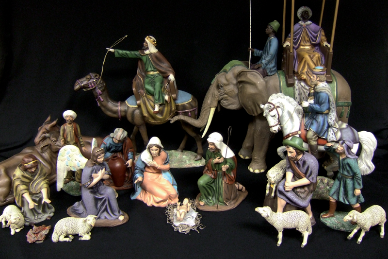 A painted clay Nativity set featuring an infant Jesus in a straw manger with the Blessed Virgin Mary and St. Joseph kneeling behind Jesus in the center. A female angel in a purple dress with white wings kneels with her arms crossed beside Mary. Two male shepherds with five white sheep, an ox and a donkey gather around the family, along with a woman presenting a jug of liquid. The three Magi ride on animals—one on a white horse, one on a camel, one on an elephant—each accompanied by an attendant.