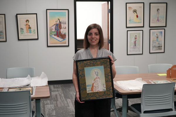 Woman standing while holding a framed artwork of an Asian Madonna. More framed Asian Madonna artworks hang on a wall behind her.