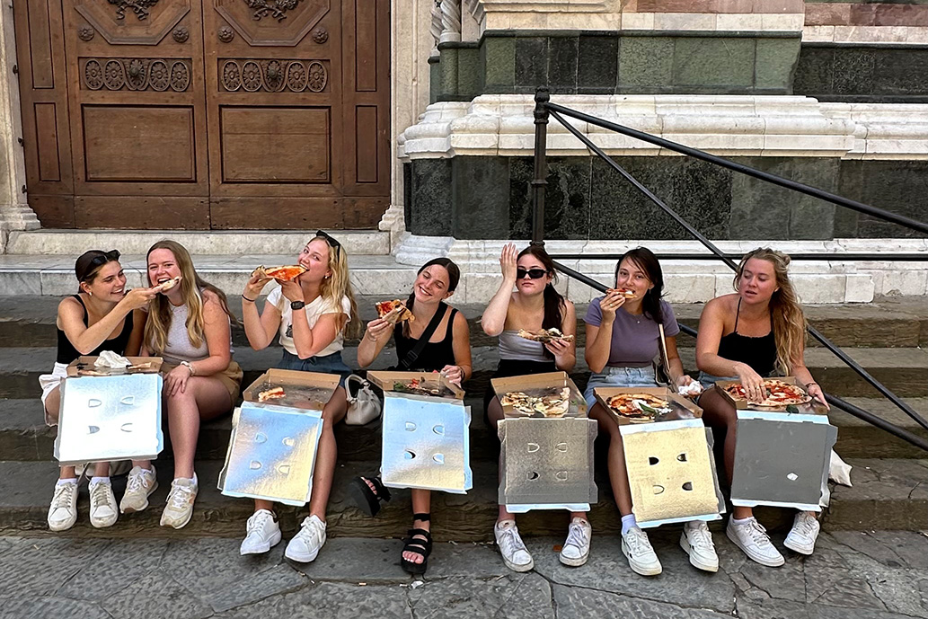Group of girls eating pizza and sitting on a curb.