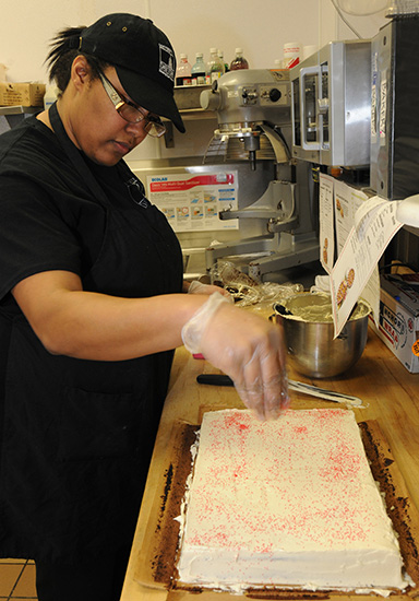 A Dining Services worker, frosts a chocolate cake with vanilla butter cream frosting and sprinkles some colorful sugar crystals on it