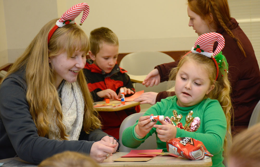 A student and child wear matching holiday hats while doing crafts.
