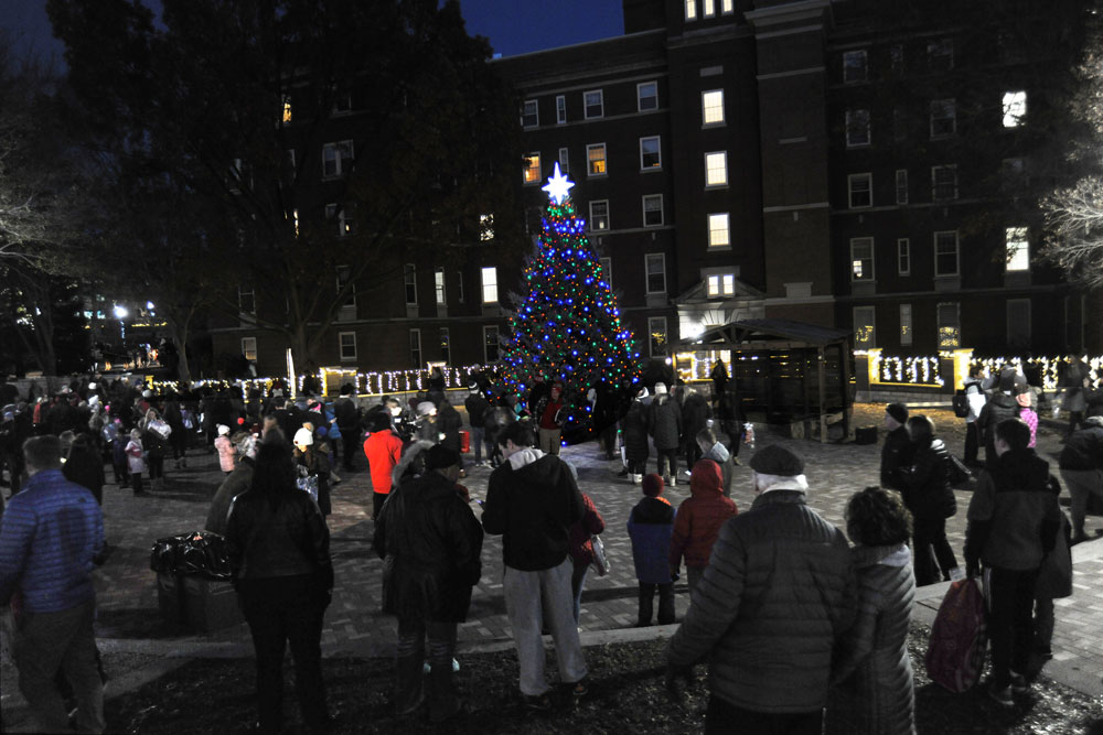A crowd is gathered around a lit Christmas tree in Humanities Plaza.