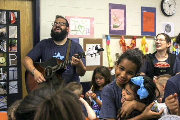 Teacher plays the guitar while students sing and hug