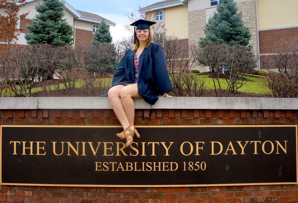 Graduate sits on the entrance sign to the University of Dayon