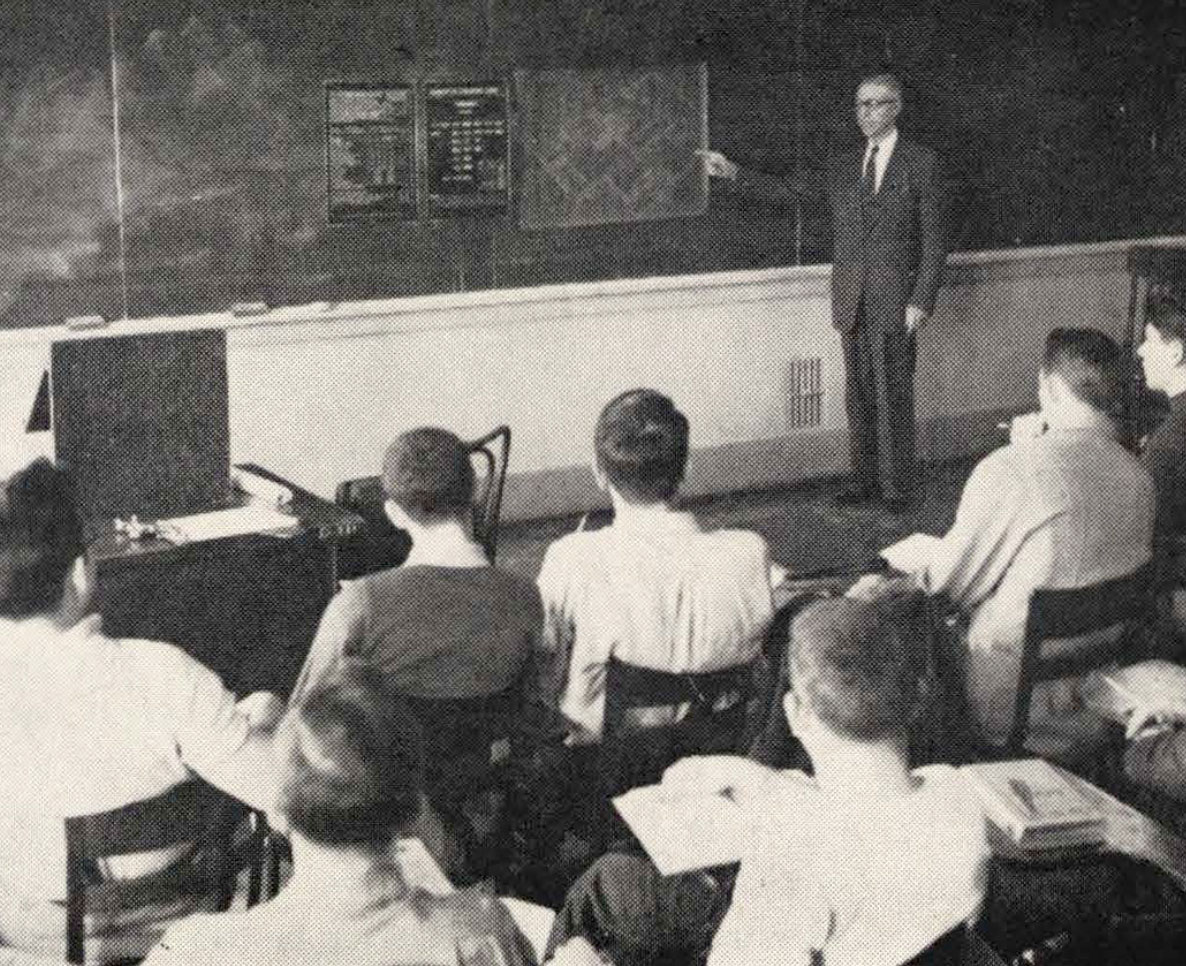Students sit at desks and watch a professor at the blackboard