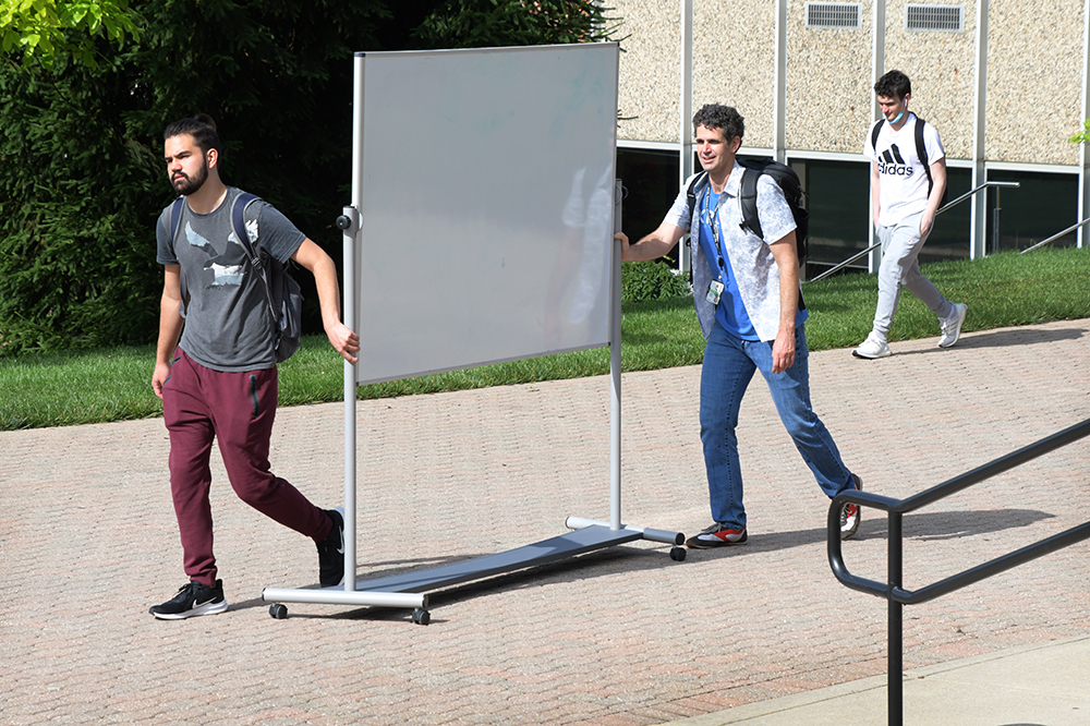 Students push a dry erase board on wheels across campus