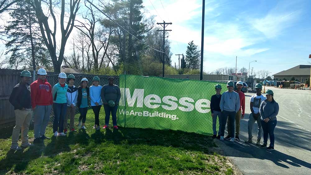 A group of students wear hard hats in front of a sign that says "Messer Construction" at a job site in Dayton.