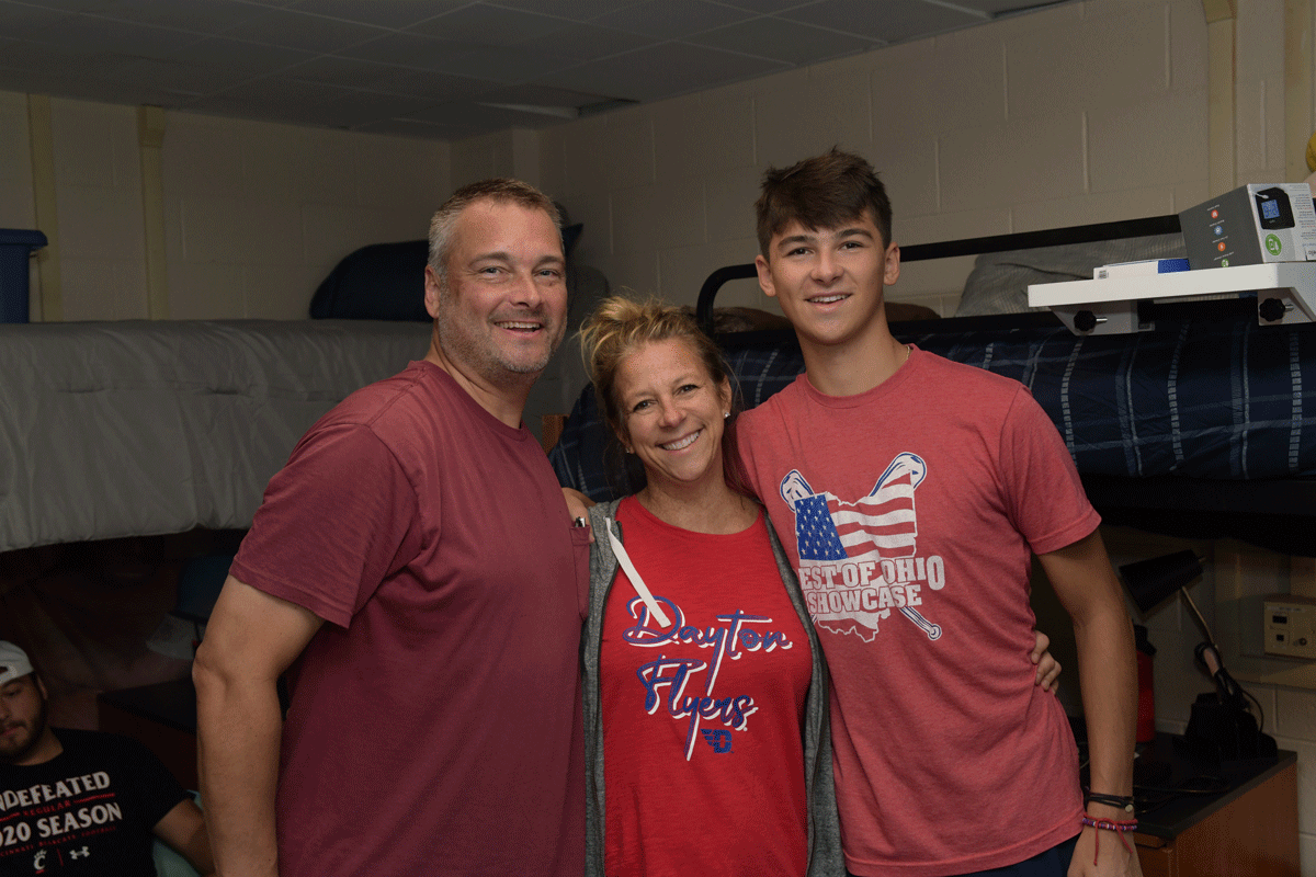 Two parents pose with their son in his dorm room, all wearing Flyer shirts.