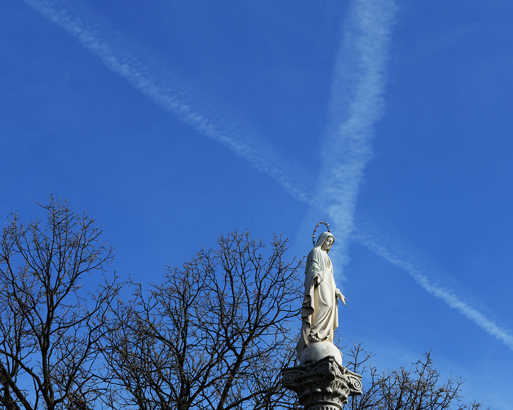 Contrails in a blue sky behind a statue of Mary.