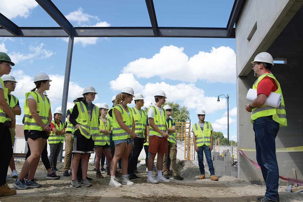 Students in hard hats and fluorescent vests gather around tour guide at a construction site.