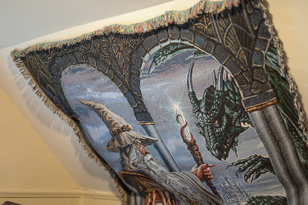 A decorative blanket hangs on a slanted ceiling depicting a wizard and a dragon.