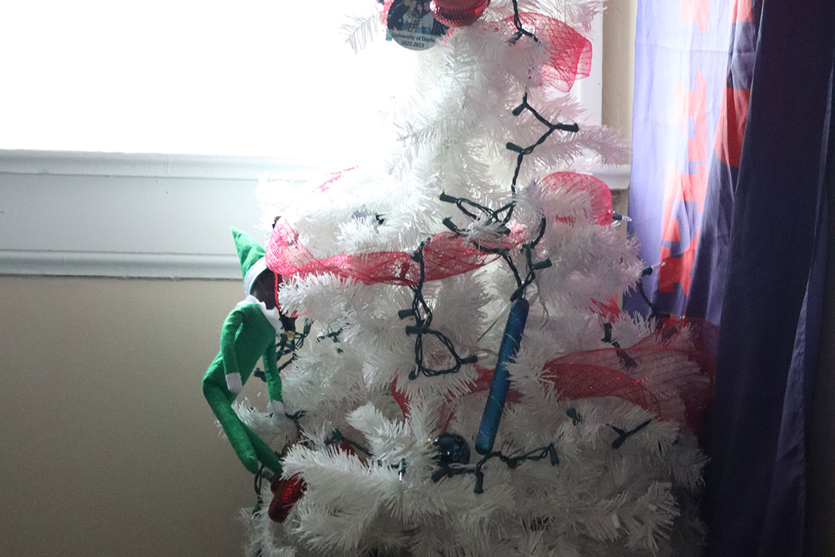 A small Elf on the Shelf clings to a small, white artificial Christmas tree.