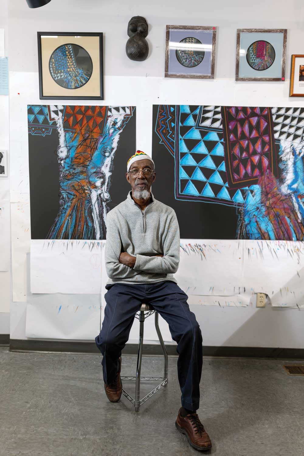 Willia "Bing" Davis poses on a stool in his studio with colorful artwork behind him.