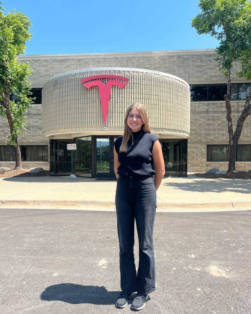 Gwendolyn stands in front of a building with the Tesla emblem on it.
