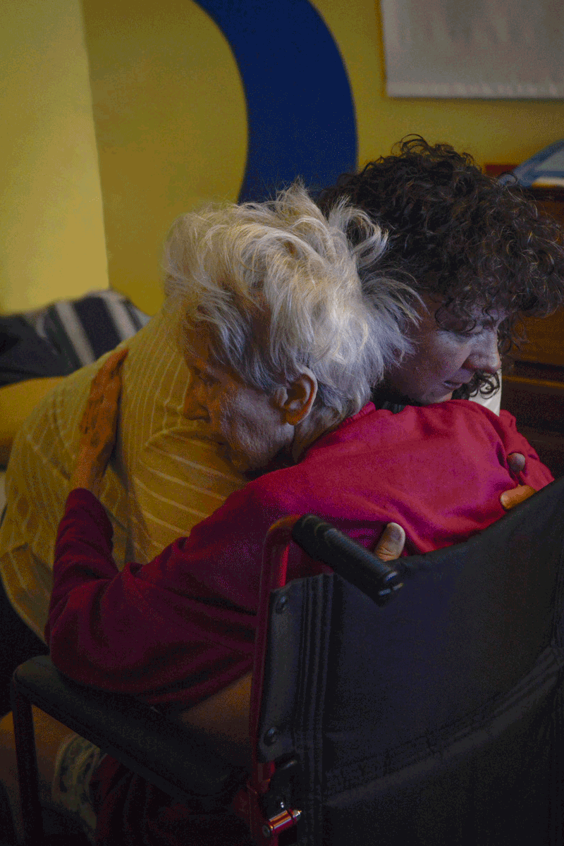 Maggie's mother lifts her grandmother from her wheelchair