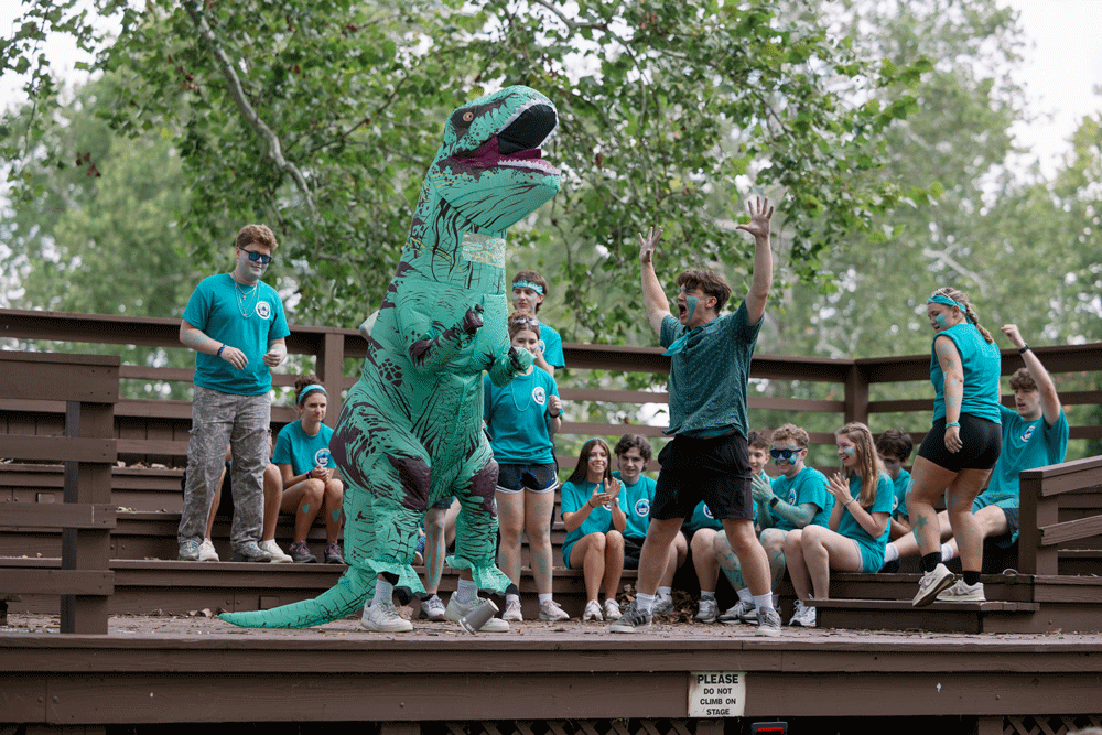 A student in an inflatable dinosaur costume puts on a skit with another group of students on a stage outside.