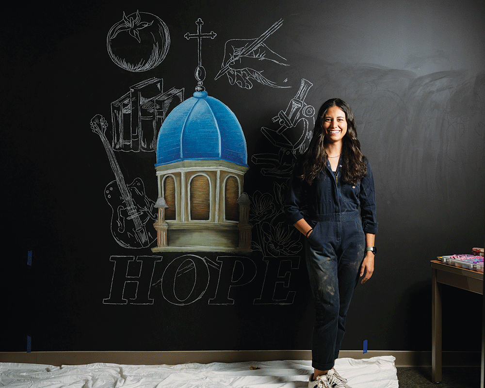 Martyn stands in front of the chalkboard where she has drawn some of the cover art including the UD chapel.