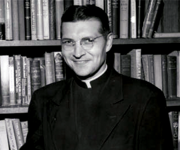 Father Philip Hoelle, S.M. smiles at the camera, wearing black and his priestly white collar, standing in front of bookshelves
