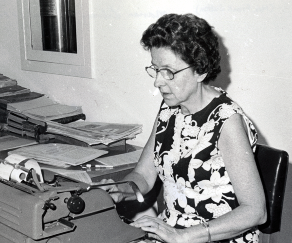 Mildred Sutton is sitting at a desk and typing on a typewriter. She is wearing glasses and a floral dress. 