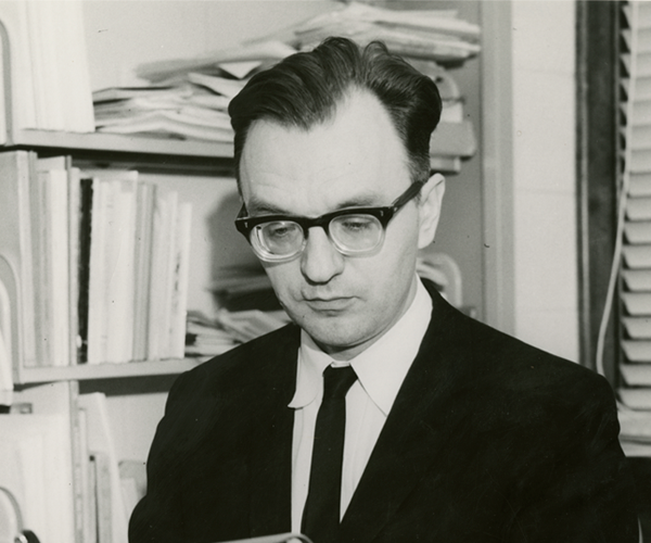 A young Brother William Fackovec, S.M. is in front of a bookcase of papers and books and looking down as if reading. He had dark hair and thick-framed glasses and is wearing a suit and tie.