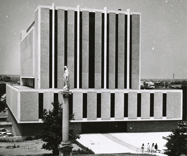Exterior view of the circa 1970-style building with a pillar and an Our Lady of the Immaculate Conception statue on top of the pillar in the foreground.