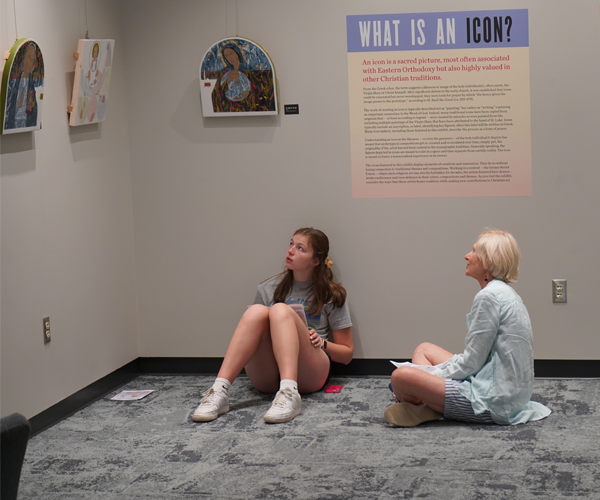 Two females sitting on the floor in a corner reflecting on the icons hung on the wall