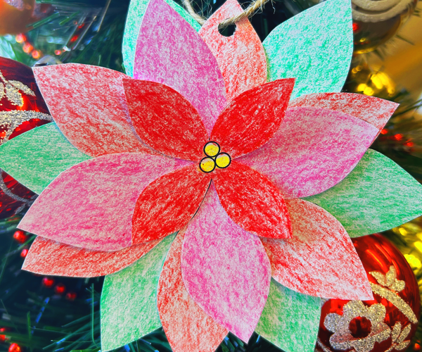 Handmade poinsettia crafted with paper and colored with crayons. It hangs amidst red and gold ornaments and pine branches.