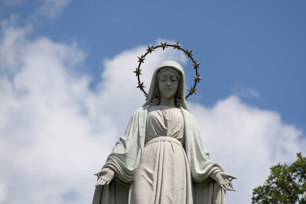Statue of Mary on UD's campus viewed form a downward angle with a blue sky, clouds and a lush treetop behind it.