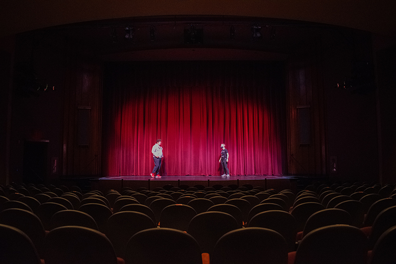 A view of the boll theatre stage from afar with two people looking at one another in front of a closed red theatre curtain