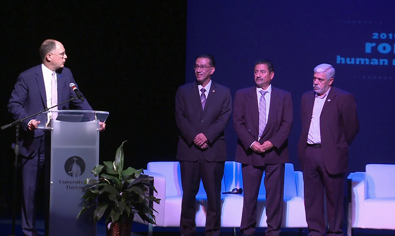 University of Dayton president, Eric Spina standing at a podium looking at three men standing on the Boll Theatre stage during the Romeo Awards